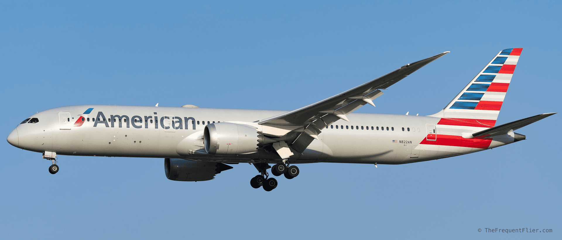 An American Airlines Boeing 787-9 in full side profile on final approach, landing gear down, in golden-hour light against a clear blue sky.