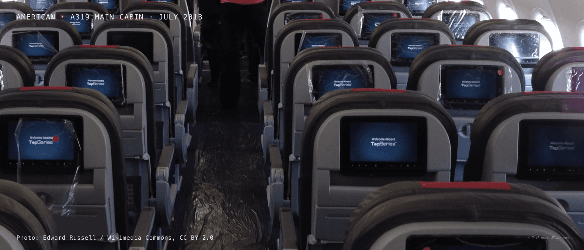 The main cabin of an American Airlines Airbus A319, rows of seatbacks receding down the aisle, each with a lit entertainment screen.