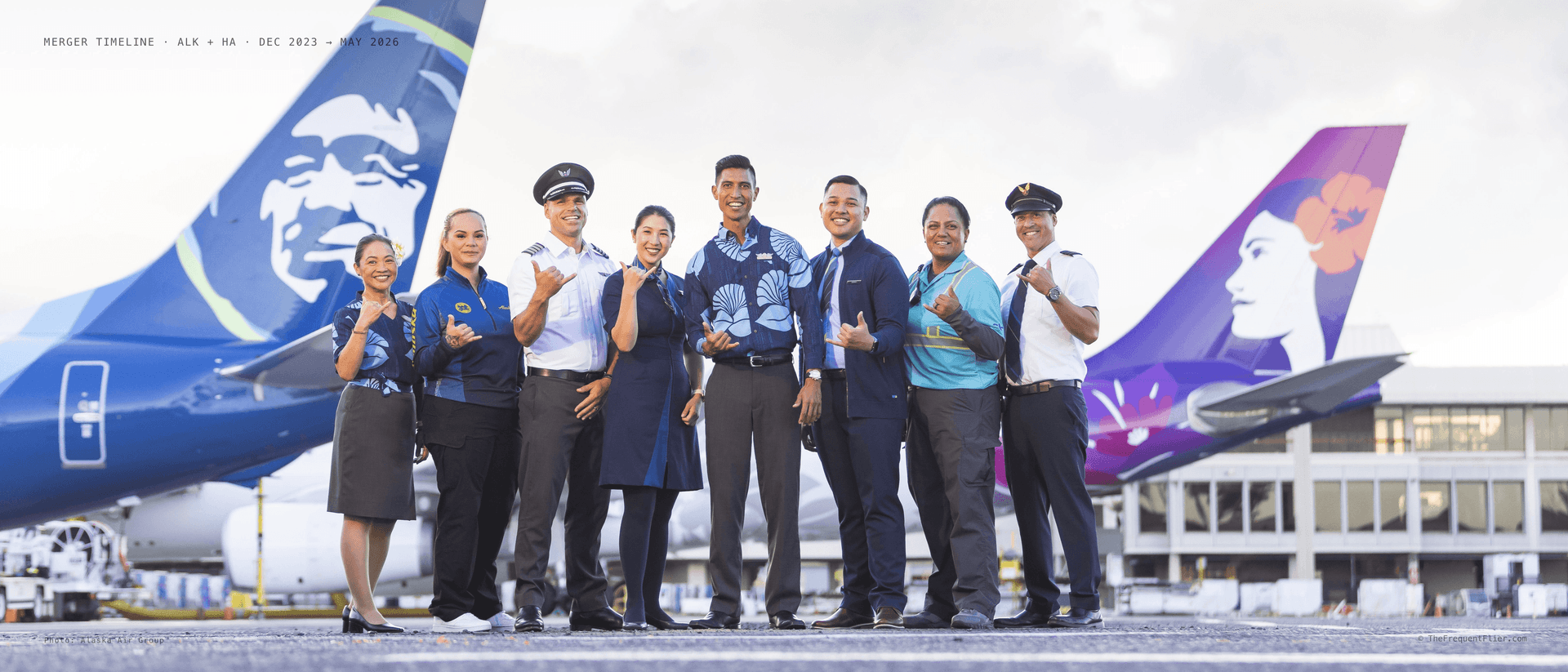 Eight Alaska and Hawaiian Airlines employees stand together on the Honolulu tarmac, framed between an Alaska 737 tail bearing the Eskimo logo on the left and a Hawaiian widebody tail bearing the Pualani logo on the right.
