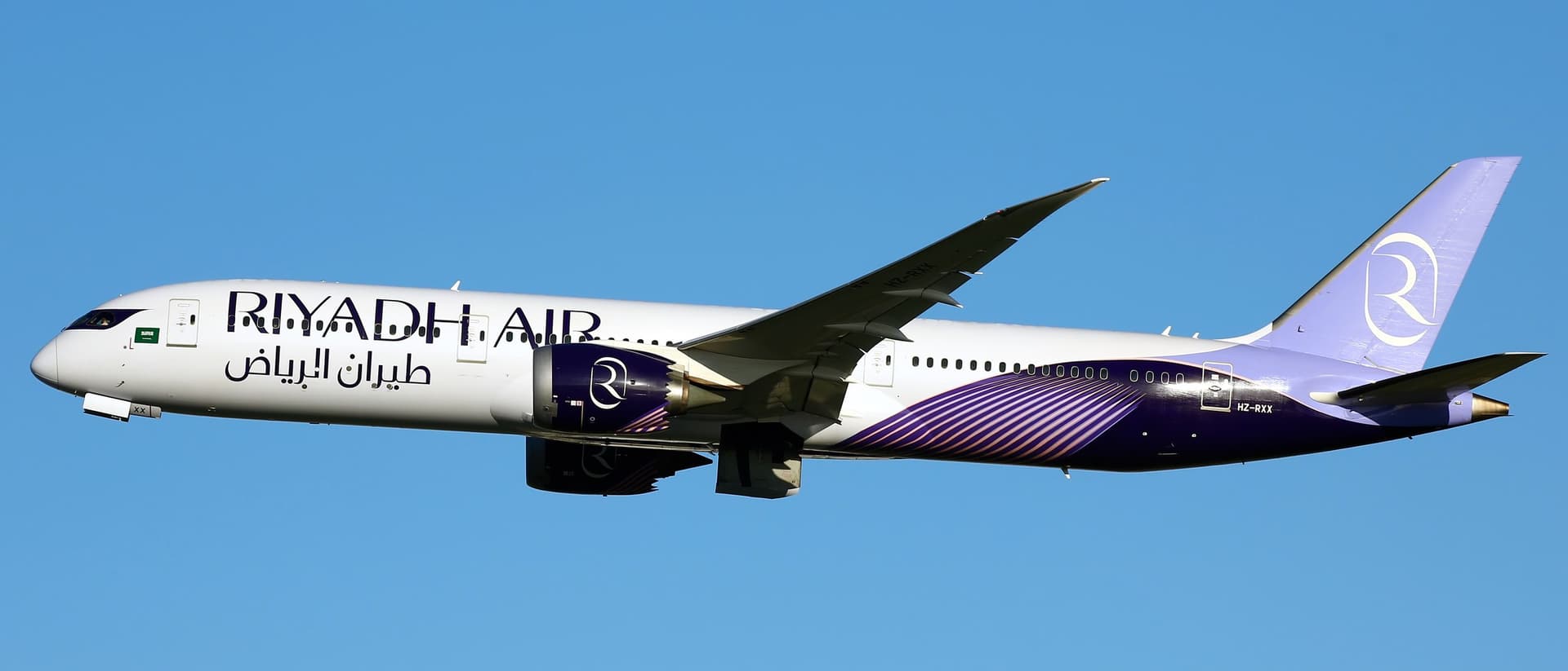 A Riyadh Air Boeing 787-9 in its deep-violet livery, banking against a clear blue sky.